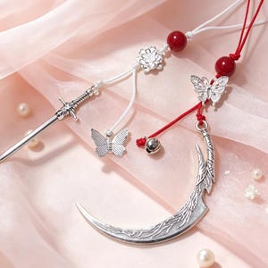 May include: Two decorative hanging ornaments featuring silver-toned metal charms. One includes a sword and a butterfly on a white cord with a red bead and flower accent. The other features a crescent moon and a butterfly on a red cord with a red bead and a small silver bell.