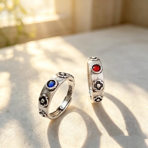Op de afbeelding: Twee zilveren ringen met bloemmotieven en edelsteenaccenten. Eén ring heeft een blauwe edelsteen, de andere een rode edelsteen. De ringen hebben een vintage stijl en zijn verstelbaar. De ringen zijn gemarkeerd met "S925".