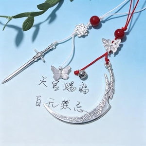 Pode incluir: Dois ornamentos decorativos suspensos com berloques prateados. Um tem um berloque de espada e borboleta num cordão branco com uma conta vermelha. O outro tem um berloque de lua crescente e borboleta num cordão vermelho com uma conta vermelha e um sino.