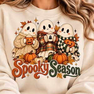 May include: A cream-coloured sweatshirt featuring a graphic of three ghosts wearing jumpers and holding mugs. The ghosts are surrounded by orange pumpkins and autumn leaves. The text "Spooky Season" is printed in orange and green cursive lettering.