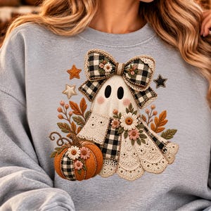 May include: A light grey sweatshirt featuring a central graphic of a ghost adorned with a black and white buffalo plaid bow, lace details, and floral accents. The design includes an orange pumpkin with plaid panels, autumn leaves, and stars.