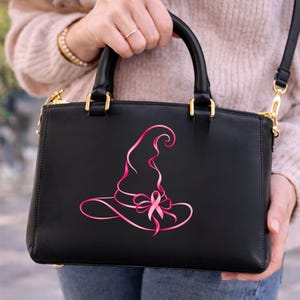 May include: A black structured handbag featuring a pink graphic design of a witch hat integrated with a breast cancer awareness ribbon. The bag has two top handles, gold-toned hardware, and a detachable shoulder strap for versatile carrying options.