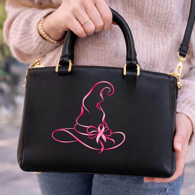May include: A black structured handbag featuring a pink graphic design of a witch hat integrated with a breast cancer awareness ribbon. The bag has two top handles, gold-toned hardware, and a detachable shoulder strap for versatile carrying options.