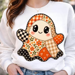 Op de afbeelding: Een wit sweatshirt met ronde hals en een grote, centrale afbeelding van een geest gemaakt van verschillende patchwork-stofpatronen. Het ontwerp van de geest bevat oranje gingham, bloemenprints, zwart-witte ruiten en stippen, allemaal omlijnd met imitatiestiksels.