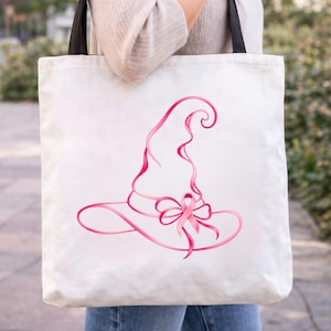 May include: A white canvas tote bag with black shoulder straps featuring a graphic illustration of a witch hat formed from a pink ribbon. The design includes a pink awareness ribbon tied into a bow at the base of the hat, centered on the bag.