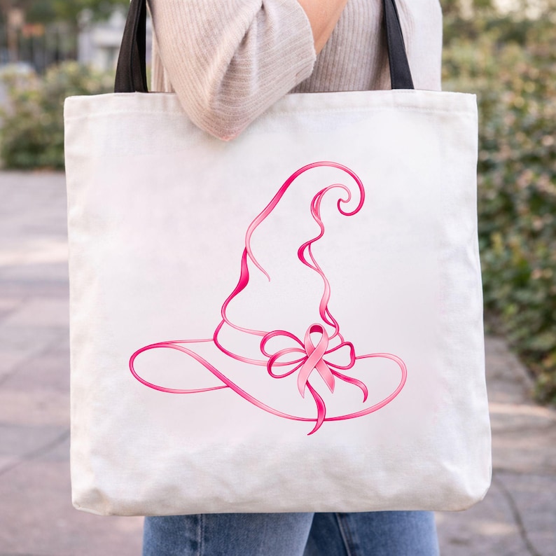 May include: A white canvas tote bag with black shoulder straps featuring a graphic illustration of a witch hat formed from a pink ribbon. The design includes a pink awareness ribbon tied into a bow at the base of the hat, centered on the bag.