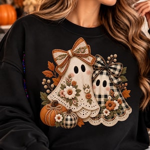 Peut inclure: Un sweat-shirt noir orné d'un motif représentant deux fantômes décorés de nœuds, de fleurs et de citrouilles. Un fantôme est crème avec un nœud texturé, tandis que l'autre présente un motif à carreaux noir et blanc. Des feuilles d'automne et des fleurs entourent les personnages.
