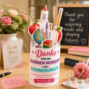 以下が含まれることがあります： ドイツ語のテキスト「Danke für die schönen Momente und Erinnerungen」が書かれた円筒形のカード。地球儀、赤いリンゴ、「You're the BEST!」と書かれた糊のボトル、鉛筆、クレヨン、ノート、積み重ねられた本のイラストが描かれています。