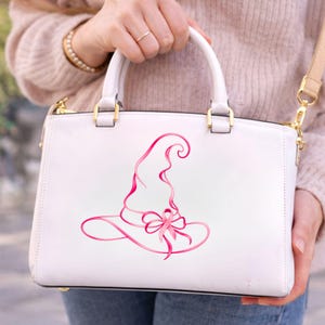 May include: A white structured handbag featuring a pink graphic illustration of a pointed witch hat adorned with a breast cancer awareness ribbon. The bag includes gold-toned metal hardware, two top handles, and a detachable shoulder strap.