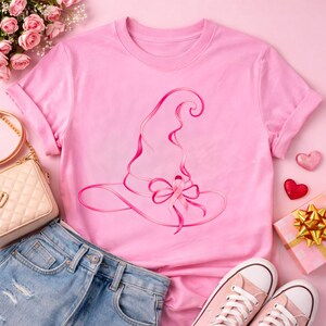 May include: A light pink short-sleeved t-shirt featuring a dark pink line art graphic of a pointed witch hat adorned with a breast cancer awareness ribbon tied into a bow at the base of the hat, centered on the front of the garment.