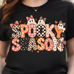 May include: A black t-shirt featuring the text SPOOKY SEASON in large, patterned letters. The design includes ghost illustrations, various bows, and motifs like leopard print, plaid, checkers, and flowers in shades of orange, pink, and black.