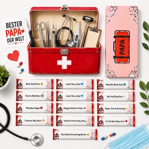 May include: A red medical kit with a white cross, filled with medical tools, and a card that says "BESTER PAPA DER WELT". Several chocolate bars with messages like "Best Dad Ever" and "Love You, Dad" are displayed.