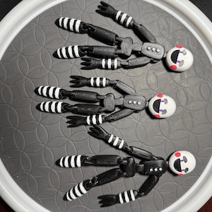 Puede incluir: Tres figuras de marionetas articuladas en blanco y negro con cabezas redondas blancas y detalles rojos están dispuestas sobre una superficie gris estampada. Las marionetas tienen extremidades articuladas con detalles de rayas blancas y negras, y están dispuestas en forma de abanico.