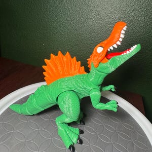 Peut inclure: Un jouet dinosaure Spinosaurus vert et orange. Le jouet a un corps vert, une voile orange et une tête orange avec des dents blanches. Le dinosaure est posé sur une surface grise.