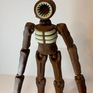 May include: A brown articulated figure with a siren-like head and a ribcage design. The figure has a circular head with a white interior, brown arms and legs, and a light-colored ribcage. The figure is a unique, detailed design.