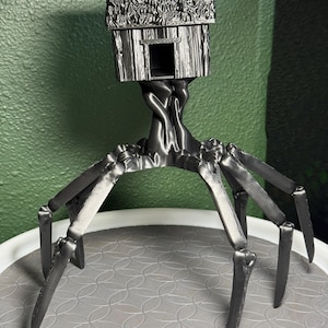 May include: A dark gray, 3D-printed sculpture of a stylized creature. It features a hut-like structure on top, supported by a tree-like trunk and multiple leg-like appendages. The sculpture is displayed on a gray, patterned surface.