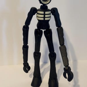 May include: A black articulated figure with a circular head featuring a white, toothed design. The figure has a skeletal torso and jointed limbs, with claw-like hands and rounded feet. The figure is a toy or collectible.