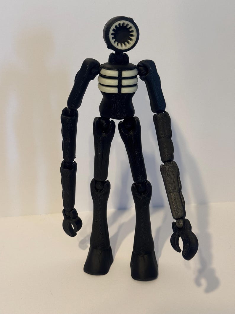 May include: A black articulated figure with a circular head featuring a white, toothed design. The figure has a skeletal torso and jointed limbs, with claw-like hands and rounded feet. The figure is a toy or collectible.