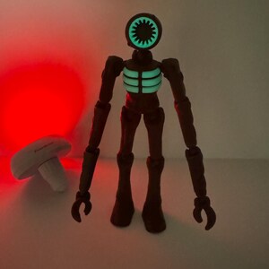 May include: A brown action figure with a siren-shaped head and glowing green details on the head and chest. The figure has articulated limbs and stands upright. A white mushroom-shaped object is in the background.