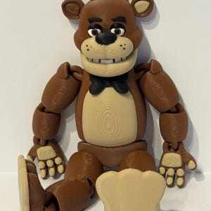 Puede incluir: Una figura articulada de Freddy Fazbear de color marrón con sombrero de copa negro y pajarita. El oso tiene un hocico y vientre color beige, con articulaciones visibles en brazos y piernas. Un micrófono de color beige está frente al oso.