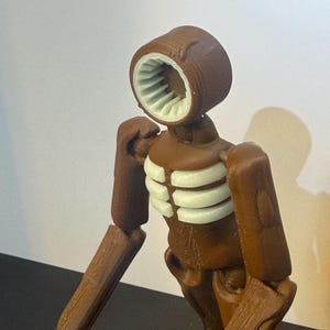 May include: A brown articulated figure with a unique head design featuring a circular opening with white interior details. The figure has a ribcage-like structure and jointed limbs, suggesting a stylized, possibly robotic, aesthetic.