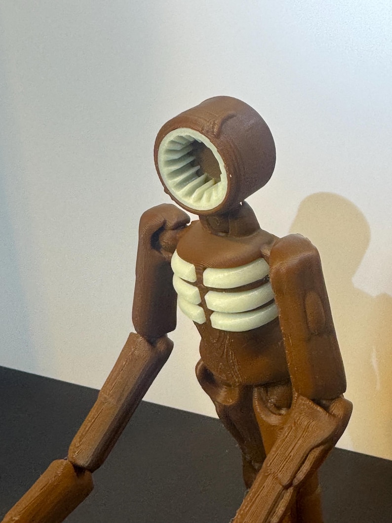 May include: A brown articulated figure with a unique head design featuring a circular opening with white interior details. The figure has a ribcage-like structure and jointed limbs, suggesting a stylized, possibly robotic, aesthetic.