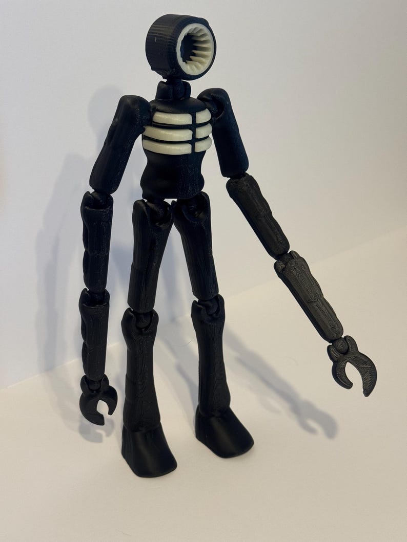 May include: A black and white articulated Siren Head figure. The figure has a cylindrical head with a speaker-like design, a white rib cage, and jointed limbs. The figure is standing on a white surface.