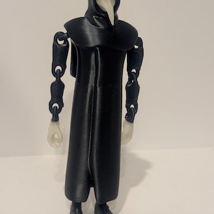 Puede incluir: Una figura de acción de médico de la peste en color negro con una túnica larga con capucha, brazos articulados y una máscara blanca con forma de pájaro. La figura tiene manos blancas y un pico blanco que contrastan con el cuerpo negro.