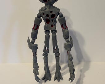 Può includere: Un action figure articolato grigio con una texture sfaccettata simile alla roccia. La figura presenta una testa rotonda e un busto coperti da fori irregolari che rivelano un interno rosso intenso. Ha arti lunghi e sottili con giunti.