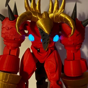 May include: A red articulated action figure with gold horns and gold drill-shaped hands. The figure features black spikes on the shoulders, a black pointed face mask, and two glowing blue circular lights on the chest area. The body has a textured surface.