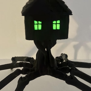 Puede incluir: Una figura articulada de color negro que presenta una pequeña estructura de casa con dos ventanas de color verde brillante, sostenida por una base central similar a un tronco que se extiende en múltiples patas largas, articuladas y arácnidas.