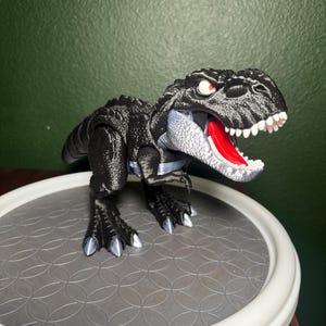 Peut inclure: Un jouet Tyrannosaurus Rex noir avec une surface texturée, des yeux rouges et une bouche rouge. Le dinosaure a des dents blanches et des griffes argentées. Il est posé sur une plateforme circulaire grise avec un bord blanc, sur fond vert.