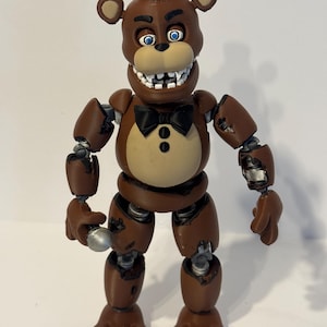 May include: A brown Freddy Fazbear action figure with a black top hat and bow tie. The figure has articulated joints and a tan chest. The bear has blue eyes and a wide, toothy grin. The figure is a collectible item.