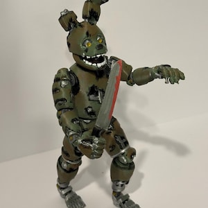 May include: A green and brown action figure of a rabbit-like character holding a bloody knife. The figure has a menacing expression with yellow eyes and a wide, toothy grin. The figure is articulated and has metallic accents.