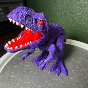 Peut inclure: Un jouet Tyrannosaurus Rex violet avec une bouche rouge et des dents blanches. Le dinosaure a des yeux rouges et est posé avec la bouche ouverte. Le jouet est posé sur une surface grise.
