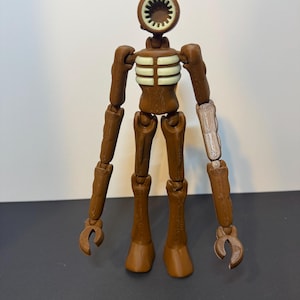 May include: A brown Siren Head action figure with a circular head and a speaker-like mouth. The figure has articulated limbs and a ribcage design. One arm is a lighter color. The figure is standing on a dark surface.
