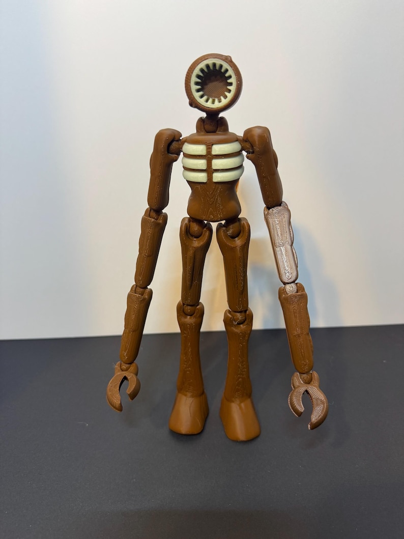 May include: A brown Siren Head action figure with a circular head and a speaker-like mouth. The figure has articulated limbs and a ribcage design. One arm is a lighter color. The figure is standing on a dark surface.
