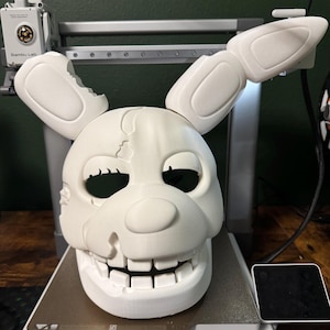 May include: A white, 3D-printed mask of a rabbit-like character with a cracked skull and open mouth. The mask has large ears and is sitting on a 3D printer bed. The Bambu Lab printer is visible in the background.