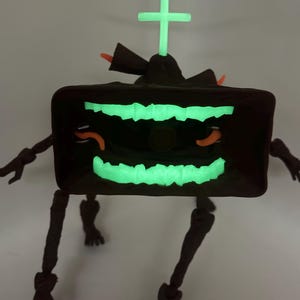 Puede incluir: Una figura robótica estilizada, de color marrón oscuro, con una cara y antena verde brillante. La figura tiene piernas y brazos articulados, y detalles en naranja. La cara tiene un diseño dentado. La antena tiene forma de cruz.