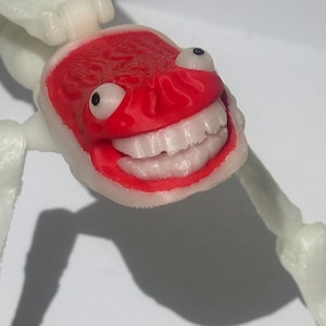May include: A 3D printed articulated figure featuring a white skeletal body and a bright red head. The head displays a wide, toothy grin with white teeth, two prominent round eyes with black pupils, and a textured surface finish throughout the piece.