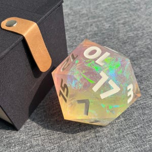 May include: A large, iridescent, twenty-sided die with white numerals. The die has a pearlescent, rainbow-like effect. A black box with a tan leather strap is in the background.