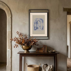 May include: A framed blue and white architectural print hangs on a grey wall. A dark wood table holds a brass bowl of dried foliage, books, and a small tray. A woven basket and a canvas tote bag sit below.
