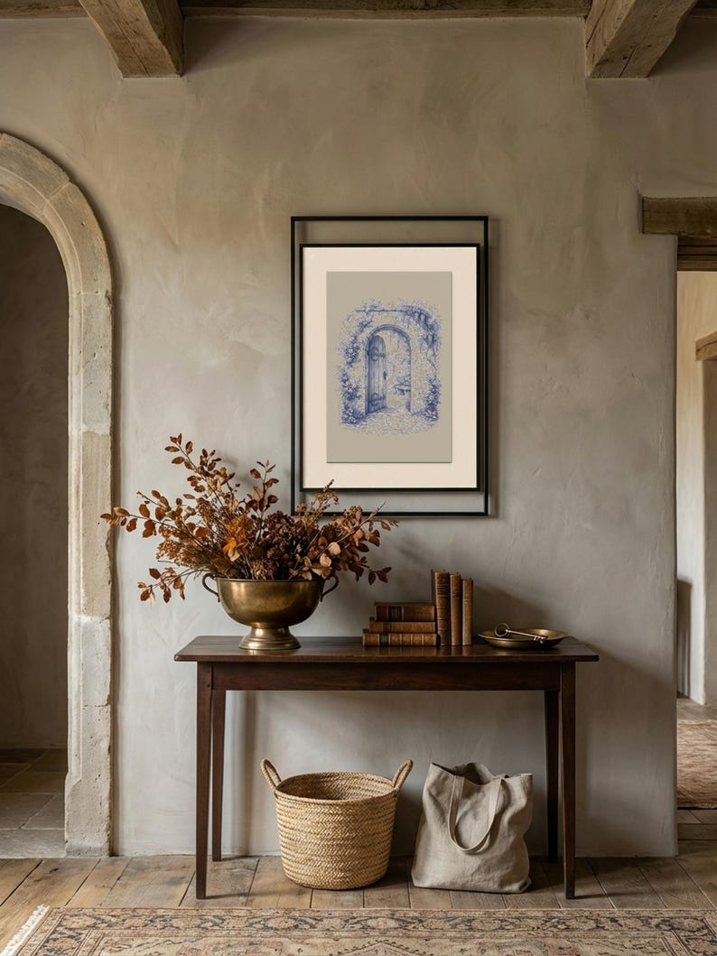 May include: A framed blue and white architectural print hangs on a grey wall. A dark wood table holds a brass bowl of dried foliage, books, and a small tray. A woven basket and a canvas tote bag sit below.