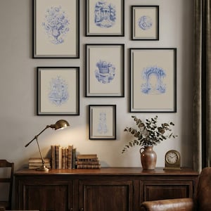 May include: A collection of eight framed blue and white prints, depicting botanical and architectural scenes. The prints are in black frames, arranged on a neutral wall. A wooden cabinet with books, a lamp, and a vase of foliage sits below the artwork.
