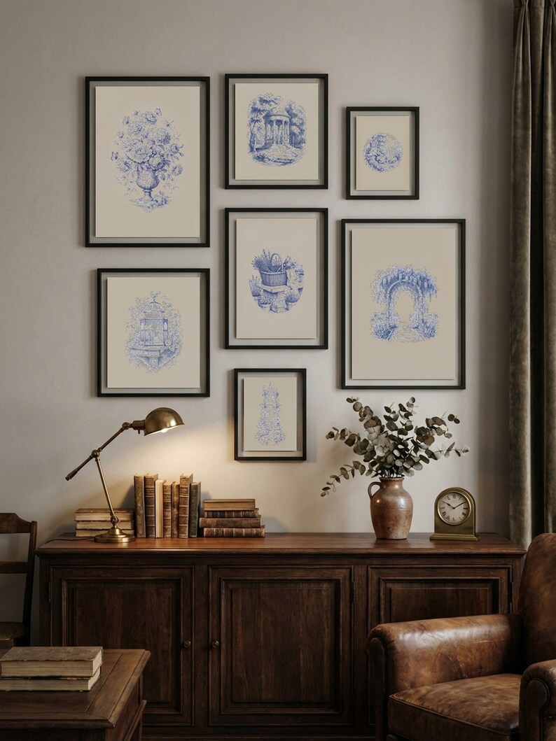 May include: A collection of eight framed blue and white prints, depicting botanical and architectural scenes. The prints are in black frames, arranged on a neutral wall. A wooden cabinet with books, a lamp, and a vase of foliage sits below the artwork.