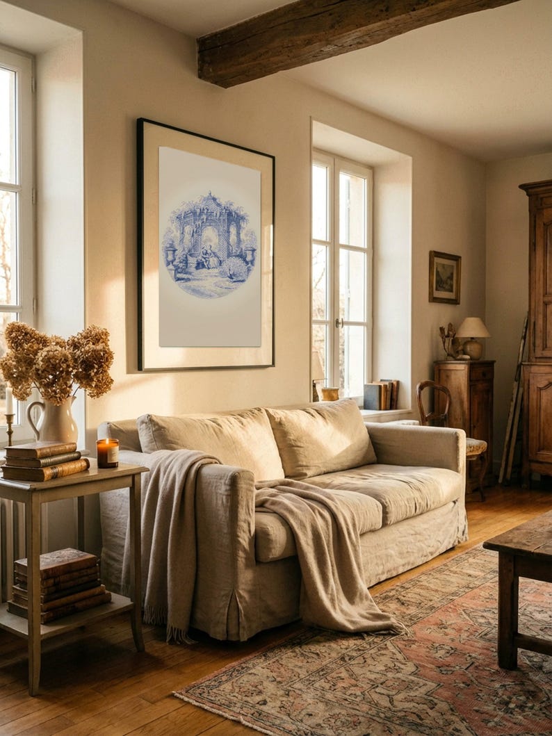 May include: A comfortable living room with a beige sofa and a throw. A framed blue and white print is on the wall. A side table displays books, dried flowers in a vase, and a candle. The room has a patterned rug and wooden flooring. The room is bathed in natural light.