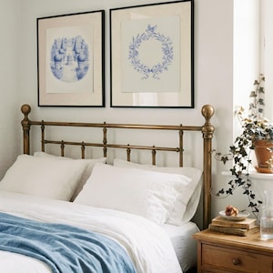 May include: A bedroom with a brass bed frame, white bedding, and a blue throw. Two framed blue and white prints are above the bed. A wooden bedside table holds books, a rose, and a glass carafe.