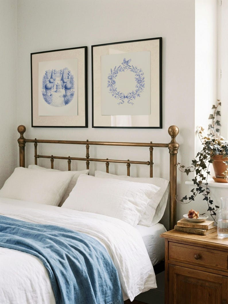 May include: A bedroom with a brass bed frame, white bedding, and a blue throw. Two framed blue and white prints are above the bed. A wooden bedside table holds books, a rose, and a glass carafe.