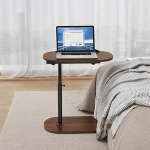 May include: A brown, adjustable laptop table with an oval tabletop and base. The table has a black, height-adjustable support column. A laptop is placed on the table, which is positioned next to a bed with a blanket.