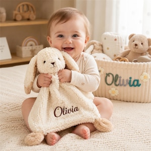 May include: A cream-coloured plush bunny security blanket featuring a soft, textured fabric and the name Olivia embroidered in dark brown thread. The blanket has long, floppy ears and small, rounded corners, providing a tactile sensory experience.